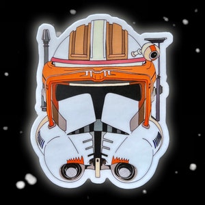 May include: A white and orange Star Wars clone trooper helmet sticker. The helmet has a white faceplate with black details and orange accents. The helmet is set against a black background with white dots.