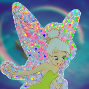 K&ouml;nnte beinhalten: Ein glitzernder, regenbogenfarbener Aufkleber von Tinkerbell, einer Fee mit gr&uuml;nen Fl&uuml;geln und einem gr&uuml;nen Kleid, vor einem blauen und violetten Hintergrund.