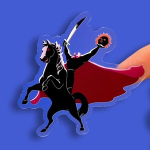 Puede incluir: Una pegatina transparente que representa a un jinete sin cabeza montado en un caballo negro encabritado. El jinete, en silueta, sostiene una espada y una calavera en llamas. La pegatina tiene una capa roja sobre un fondo azul.