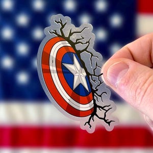 May include: A clear sticker with a cracked design, featuring Captain America's shield with red, white, and blue colors. The shield is partially obscured by the cracked design, creating a dramatic effect.