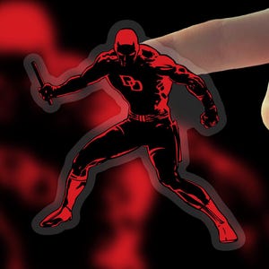 May include: A red and black sticker featuring a comic book character in a dynamic pose, holding a baton. The character is wearing a red suit with a stylized "D" logo. The background is black with red accents.