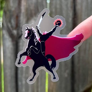 Puede incluir: Una pegatina que representa a un jinete sin cabeza montado en un caballo negro encabritado. El jinete empu&ntilde;a una espada y una calavera en llamas. La pegatina tiene una capa roja y rosa y un contorno plateado. El dise&ntilde;o est&aacute; inspirado en el cuento cl&aacute;sico.