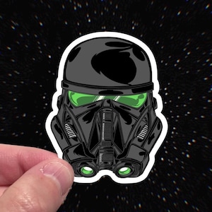 May include: A black and green sticker featuring a stylized illustration of a Death Trooper helmet from Star Wars. The helmet has green lenses and is set against a black background with white stars.