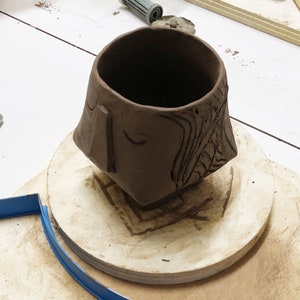 May include: A handmade brown clay mug with carved details. The mug features a square base and a rounded top. One side has a rectangular shape and a curved line, while the other side has carved lines. The mug sits on a wooden surface.