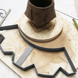 May include: A brown clay pot sits on a wooden base, centered within a gray plastic mold with a scalloped edge. The mold is on a wooden surface.