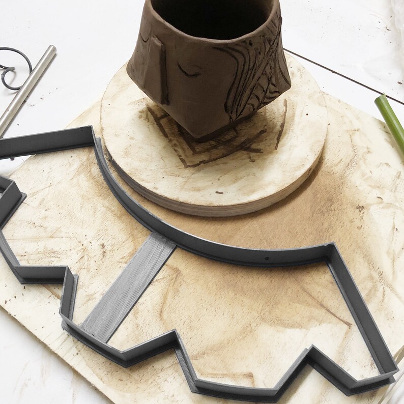 May include: A brown clay pot sits on a wooden base, centered within a gray plastic mold with a scalloped edge. The mold is on a wooden surface.