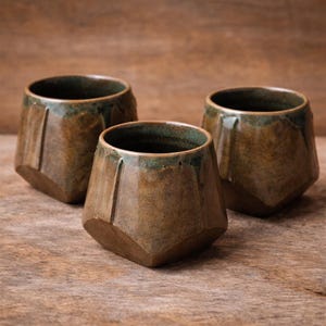 Może przedstawiać: Trzy ręcznie robione ceramiczne kubki o fasetowym wzorze. Kubki są mieszanką brązu i zieleni z błyszczącym wykończeniem. Kubki są prezentowane na drewnianej powierzchni.