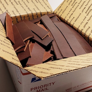 May include: A cardboard box filled with brown leather strips. The box is labeled "Priority Mail" and has the USPS logo.