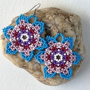 Peut inclure: Une paire de boucles d'oreilles en forme de fleur, composées de perles bleues, roses, blanches et violettes. Les boucles d'oreilles sont suspendues à des crochets argentés.