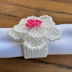 Peut inclure: Un rond de serviette blanc perlé en forme de fleur avec un centre rose. La bague est faite de petites perles rondes et repose sur un tissu blanc. Le motif floral ajoute une touche décorative à la table.