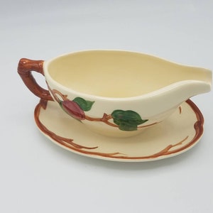 May include: A white ceramic gravy boat with a brown twig handle and a matching saucer. The boat and saucer are decorated with a brown twig design and green leaves with red berries.