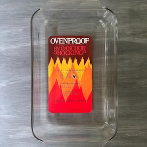 May include: A clear, rectangular glass baking dish with rounded corners. The dish has a red and orange label that reads "OVENPROOF BY ANCHOR HOCKING" and "1 BAKING DISH 2 QT/1.9L". The dish is designed for oven use.