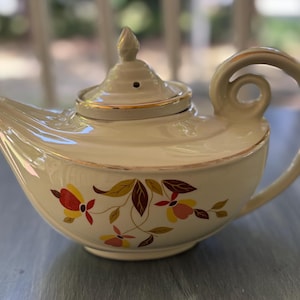 May include: Cream-colored ceramic teapot with a curved handle and spout. The teapot features a floral design in red, yellow, and brown. Gold trim accents the lid and rim, adding a touch of elegance to this vintage-style tea pot.