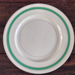 May include: A white ceramic plate with a green band around the rim and a gold trim. The plate has a simple, elegant design, suitable for serving meals. The plate is round and appears to be vintage.