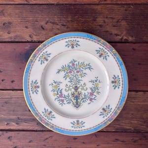 May include: A white porcelain plate with a gold rim and a light blue border. The plate features a central floral design with pink, yellow, and blue flowers. Additional floral motifs are placed around the border.