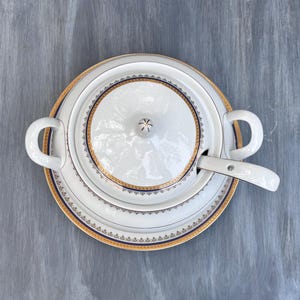 May include: White porcelain soup tureen with a lid, handles, and a matching spoon. The tureen and spoon have gold and navy blue trim. The tureen sits on a matching plate. The background is a gray wooden surface.