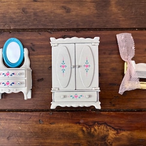 May include: Miniature doll furniture set including a white dresser with a blue mirror, a white armoire, and a gold-colored crib with a pink net. The furniture is decorated with pink and teal floral accents.