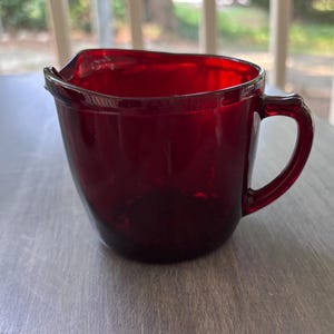 May include: A ruby red glass creamer with a handle and a pouring spout. The creamer is made of a transparent glass material and has a rounded shape. The handle is arched and attached to the side of the creamer.