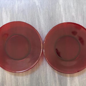 May include: Two matching red glass saucers. Each saucer is round with a slightly raised rim and a recessed center. The color is a deep, rich red, and the glass appears to be smooth and glossy. The saucers are placed side by side.
