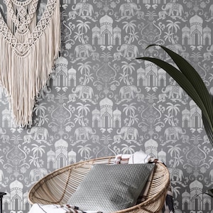 May include: A grey and white patterned wallpaper with a repeating design of elephants, palm trees, and ornate buildings. A wicker chair with a grey cushion and a throw blanket sits in front of the wallpaper. A small black side table with a vase of greenery sits next to the chair.