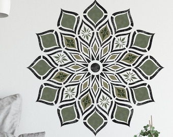 May include: A grey sofa with cushions and a throw blanket in front of a white wall with a large green and black mandala stencil design. A small potted plant sits on a brown side table.
