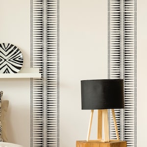 Könnte beinhalten: Ein Raum mit cremefarbener Wand mit zwei vertikalen schwarz-weißen Streifen. Eine schwarze Lampe mit Holzfuß steht auf einem Holzblock. Ein schwarz-weiß gemusterter Teller liegt auf einem weißen Regal.