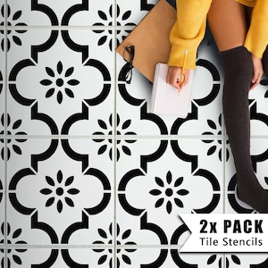 Può includere: Due stencil per piastrelle in bianco e nero con un motivo geometrico di fiori e linee curve. Gli stencil sono su un pavimento bianco e una persona è seduta sul pavimento con un taccuino e una penna.