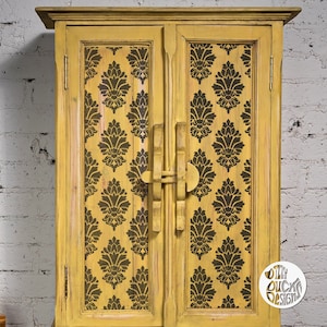 Può includere: Un armadio in legno giallo con due ante. Le ante sono decorate con un motivo damascato bianco e nero. L'armadio ha un design semplice con un unico chiavistello al centro.