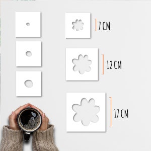 May include: White square stencils with flower cutouts in three sizes: 7 cm, 12 cm, and 17 cm. Three smaller squares show the progression of the flower's center. A person holds a black mug of coffee.