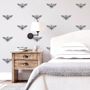 Op de afbeelding: Een witte slaapkamer met een bed, nachtkastje en lamp. De muur is versierd met een patroon van grijze bijensilhouetten.