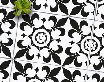 May include: Overhead shot of white tiles with a black fleur-de-lis and floral pattern. A small green succulent plant in a white pot sits in the upper left corner. The tiles are arranged in a grid.