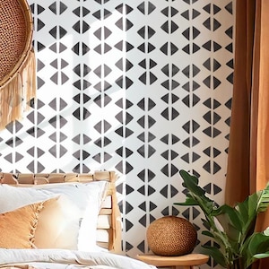 May include: A bedroom scene featuring a white wall with a repeating black geometric pattern. A woven headboard and pillows are visible, along with a small wooden side table holding a woven sphere. Brown curtains and a hanging woven basket add warmth.