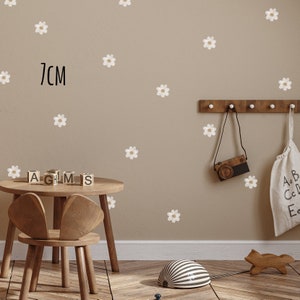 May include: A child's room with daisy wall stickers, a small wooden table and chair, and decorative items. The wall features white daisy stickers, with the text "7cm" in black. A wooden camera and a bag hang on the wall.