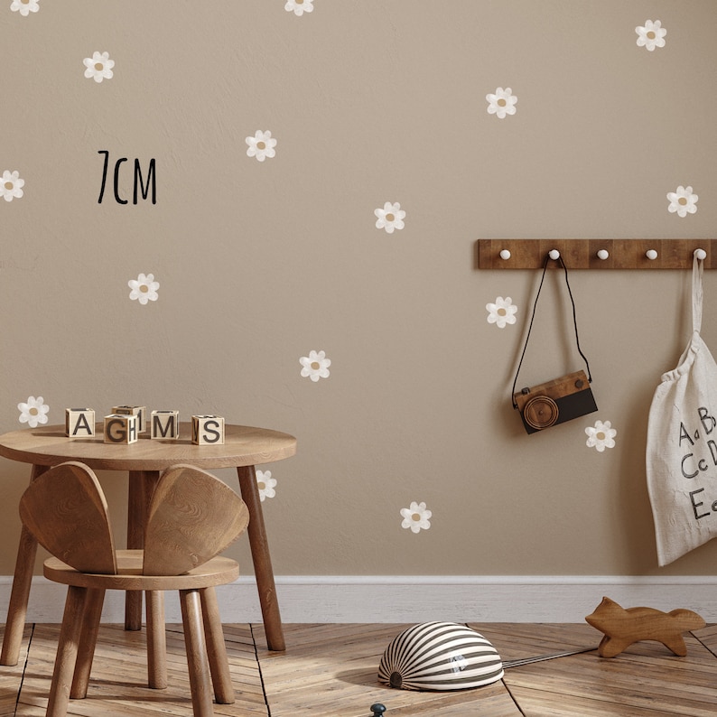 May include: A child's room with daisy wall stickers, a small wooden table and chair, and decorative items. The wall features white daisy stickers, with the text "7cm" in black. A wooden camera and a bag hang on the wall.