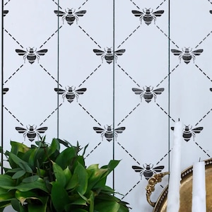 Op de afbeelding: Witte muur met een zwart bijenstencilpatroon. De bijen zijn gerangschikt in een ruitvormig patroon met een gestippelde lijn die ze verbindt. De muur is wit geschilderd en de bijen zijn zwart. Er zijn wat groene planten in de linkerbenedenhoek van de afbeelding.