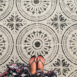 Könnte beinhalten: Draufsicht auf Füße mit schwarzen Flip-Flops und rotem Nagellack, die auf einem gemusterten Fliesenboden stehen. Die Fliesen sind weiß mit einem schwarzen Kreis- und Blumendesign.