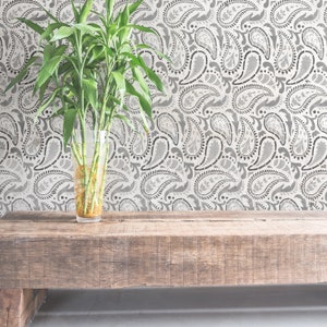 May include: A wooden bench with a vase of green bamboo plants in front of a wall with a grey paisley pattern.