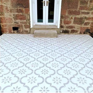 Può includere: Pavimento piastrellato bianco e grigio con un motivo floreale. Le piastrelle sono disposte in un motivo ripetuto, creando una superficie decorativa. Il pavimento è di fronte a una porta bianca con maniglie dorate e un muro di mattoni.