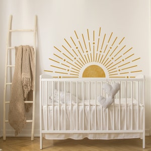 May include: A white cot with a white cot sheet and two grey heart-shaped pillows. The wall behind the cot has a gold sunburst decal with a yellow sun.