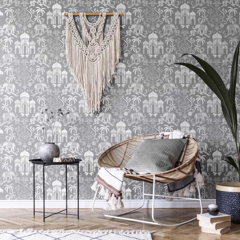 May include: A grey and white patterned wallpaper with a repeating design of elephants, palm trees, and ornate buildings. A wicker chair with a grey cushion and a throw blanket sits in front of the wallpaper. A small black side table with a vase of greenery sits next to the chair.