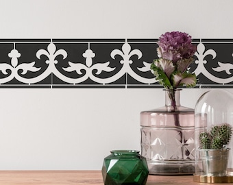 May include: A black and white tile border with a decorative pattern. The border is displayed on a white wall with a vase of purple flowers, a green glass jar, and a glass cloche with a cactus plant inside.