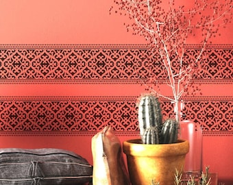 May include: Two black stencil designs on a red wall, one above the other. The designs are intricate and feature a repeating pattern of geometric shapes. A potted cactus and a brown leather bag are in the foreground.