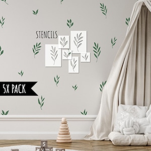 Op de afbeelding: Een grijze muur versierd met witte bladstencils en groene bladstickers. De stencils zijn gerangschikt in een groep van vijf, met het woord "STENCILS" erboven. Een zwarte banner leest "5X PACK". Een houten stapelspeelgoed en blokken staan op de voorgrond.