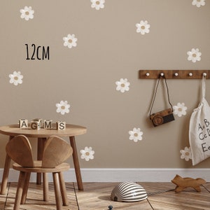 May include: A beige wall adorned with white and tan daisy decals. The text "12cm" is visible. A small wooden table and chair are in the foreground. A wooden rack holds a toy camera and a bag with letters.
