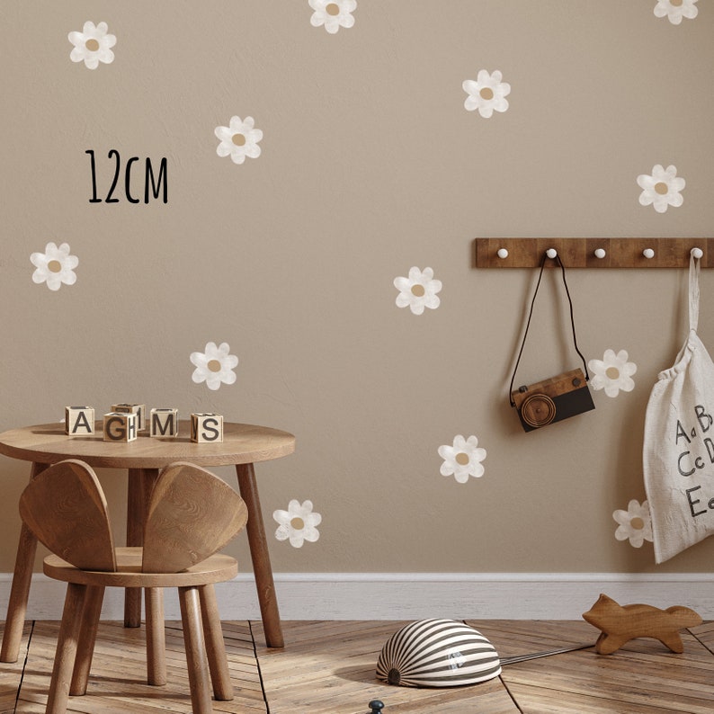 May include: A beige wall adorned with white and tan daisy decals. The text "12cm" is visible. A small wooden table and chair are in the foreground. A wooden rack holds a toy camera and a bag with letters.