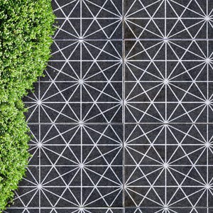 May include: Overhead view of a geometric tile pattern in black and white. The tiles are arranged in a grid, with each tile featuring a starburst design. A vibrant green hedge borders the left side of the image.
