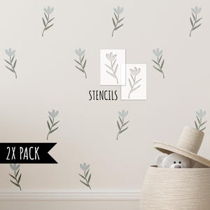 Op de afbeelding: Een lichtbeige muur versierd met een bloemmotief van blauwgrijze bloemen en groene stelen. Twee witte sjablonen met hetzelfde bloemdesign worden weergegeven met het woord "STENCILS" eronder. Een zwarte pijl leest "2X PACK".