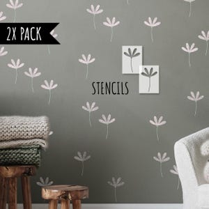 Op de afbeelding: Een grijze muur versierd met lichtroze bloemstencils. Het woord "STENCILS" is in het zwart gedrukt. Een zwarte banner leest "2X PACK". Een stapel dekens en een houten kruk staan op de voorgrond.
