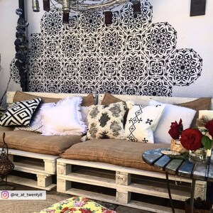 May include: A white pallet couch with brown cushions and pillows. The couch is decorated with black and white patterned pillows, a white pillow with fringe, and a white pillow with black embroidery. There are red roses in a glass vase on a small wooden table next to the couch. The wall behind the couch is decorated with a black and white stencil design.