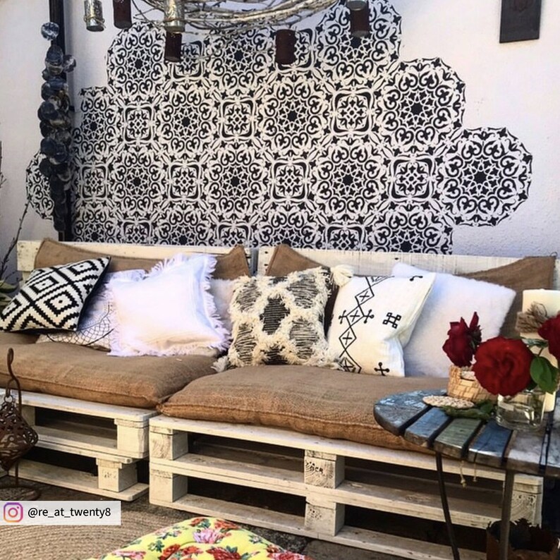 May include: A white pallet couch with brown cushions and pillows. The couch is decorated with black and white patterned pillows, a white pillow with fringe, and a white pillow with black embroidery. There are red roses in a glass vase on a small wooden table next to the couch. The wall behind the couch is decorated with a black and white stencil design.
